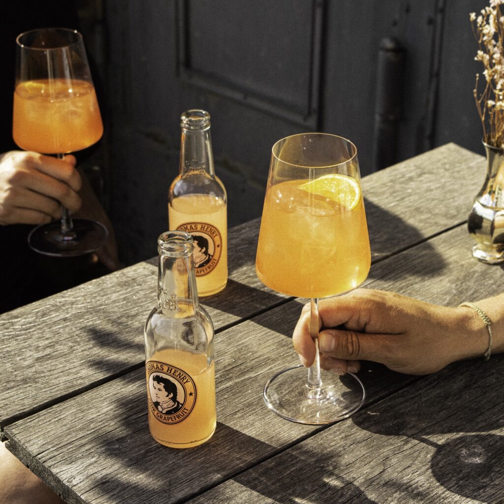 Zwei Weingläser mit Aperol Grapefruit Spritz und Eis, daneben zwei Flaschen Thomas Henry Pink Grapefruit auf einem Holztisch im Sonnenlicht