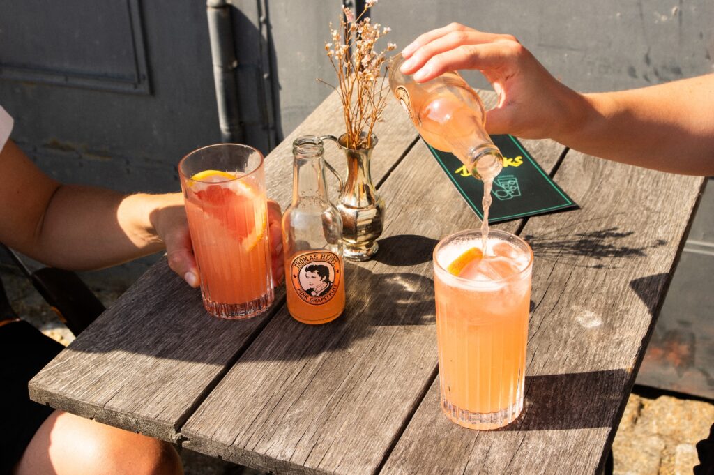 Thomas Henry Pink Grapefruit wird in ein Longdrinkglas mit Eis gegossen, daneben ein zweiter Tequila-Cocktail mit Grapefruitscheibe auf sonnigem Holztisch