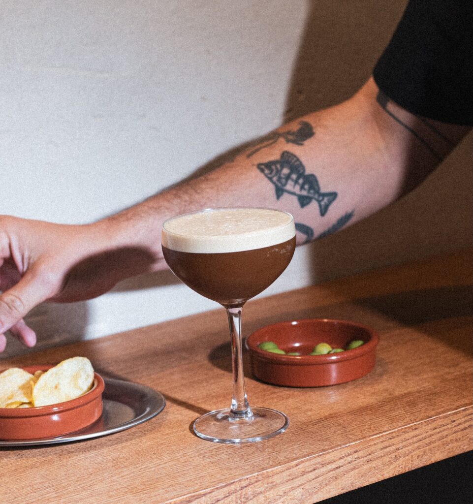 Espresso Martini in der Coupette mit cremiger Schaumkrone auf einem Holztresen, daneben Chips und ein Schälchen mit Oliven